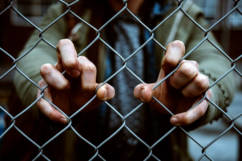 Teenage Juvenile wearing a green hoodie with their fingers wrapped around the links of a chain link fence